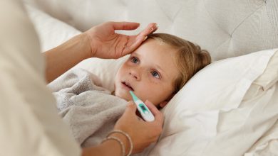 Yatakta yatan küçük bir çocuğun ateşi ölçülüyor. Bir yetişkin elini çocuğun alnına koyarken, diğer eliyle ağızdan ateş ölçer tutuyor. Çocuk halsiz ve hasta görünüyor; sahne ev ortamında bir hastalık durumunu yansıtıyor.