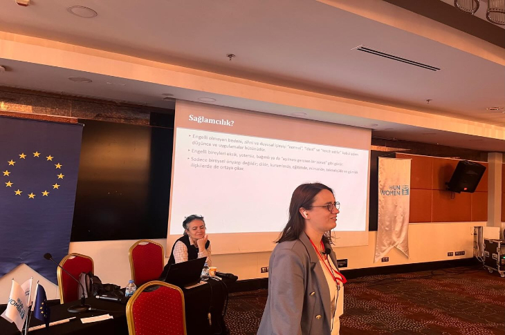 Bir konferans salonunda düzenlenen oturumda, projeksiyon ekranında “Sağlamcılık?” başlıklı bir sunum yer alıyor. Ön planda konuşmacılardan biri ayakta, arkada ise dizüstü bilgisayarıyla oturmuş başka bir katılımcı bulunuyor. Solda Avrupa Birliği bayrağı, sağda ise UN Women logosu görülüyor.