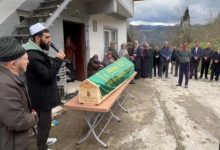 Bir imamın dua ettiği, etrafında insanların toplandığı ve yeşil örtüyle kaplı bir tabutun önünde gerçekleştirilen bir cenaze töreni görülüyor.