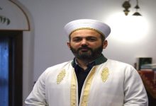 Geleneksel dini kıyafetler giymiş bir imam, kapalı bir mekânda kameraya doğru bakarak duruyor. Üzerinde beyaz cübbe ve sarık bulunuyor; arka planda ise cami içini andıran sade bir ortam görülüyor.