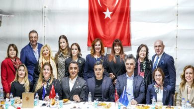 Fotoğrafta kapalı bir salonda düzenlenen bir iftar programında toplu halde poz veren bir grup görülüyor. Masanın önünde oturan ve arkada ayakta duran kadın ve erkeklerden oluşan kalabalık bir grup bulunuyor. Masanın üzerinde yemek tabakları, su şişeleri ve küçük bayraklar yer alıyor. Arka duvarda büyük bir Türk bayrağı asılı. Katılımcılar resmi kıyafetlerle kamera karşısında birlikte hatıra fotoğrafı çektiriyor.