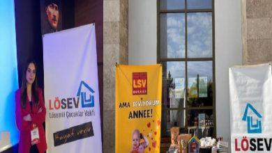 Fotoğrafta kapalı bir etkinlik alanında kurulan bir tanıtım standı görülüyor. Solda kırmızı ceketli bir kadın, LÖSEV (Lösemili Çocuklar Vakfı) logosunun yer aldığı bir afişin yanında duruyor. Ortada sarı renkte “LSV Dükkan – Ama seviyorum anne!” yazılı bir banner ve önünde çeşitli ürünlerin bulunduğu bir masa yer alıyor. Sağ tarafta ise yine LÖSEV logosunun bulunduğu başka bir afiş bulunuyor. Arka planda yüksek bir duvar, büyük bir pencere ve duvarda asılı bir portre görülüyor.