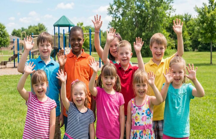 Bir parkta çekilen görüntüde, farklı çocuklar yan yana durup ellerini kaldırarak gülümsüyor. Renkli kıyafetler içindeki grup, neşe, birlik ve kapsayıcılık hissini yansıtıyor.