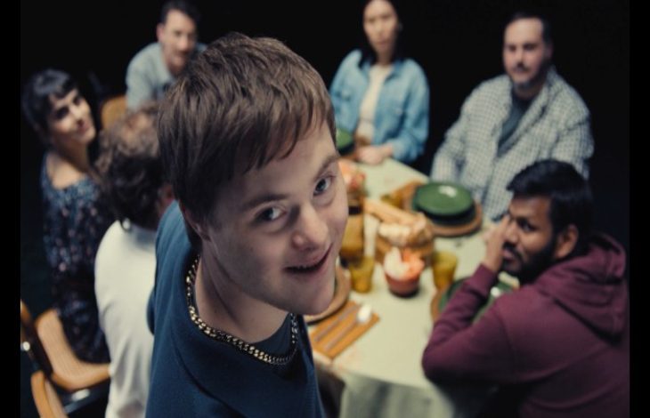 Bir yemek masasının etrafında toplanmış bir grup insan görülüyor. Ön planda, kameraya dönüp gülümseyen genç bir birey dikkat çekerken, masadaki diğer kişiler ona bakıyor. Ortam samimi bir aile ya da arkadaş buluşmasını andırıyor.