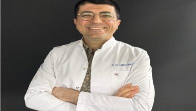 Fotoğrafta beyaz önlük giymiş bir diş hekimi görülüyor. Kollarını göğsünde birleştirmiş şekilde duruyor ve kameraya gülümseyerek poz vermiş. Yuvarlak çerçeveli gözlük takıyor. Arka plan koyu renk olduğu için önlük ve yüzü daha belirgin şekilde öne çıkıyor.