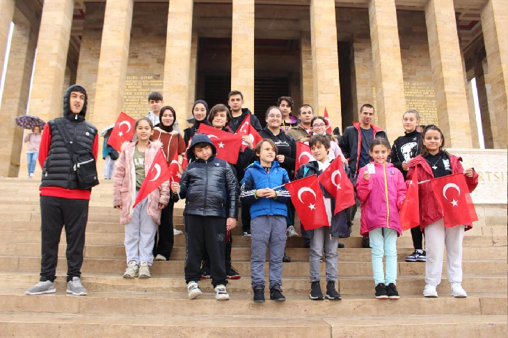 Görselde, Anıtkabir’in merdivenlerinde bir araya gelmiş çocuklar ve gençlerden oluşan bir grup görülüyor. Çoğu elinde Türk bayrağı taşıyor. Yağışlı havaya uygun mont ve yağmurluklar giyen grup, anıtsal yapının sütunları önünde hatıra fotoğrafı veriyor.