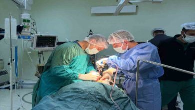 Bir ameliyathanede çekilen fotoğrafta, cerrahi kıyafet ve maskeler takan iki sağlık görevlisi ameliyat masasında yatan hastaya müdahale ediyor. Etraflarında ameliyat lambaları, tıbbi cihazlar ve monitörler bulunurken arka planda başka sağlık personeli de görülüyor.