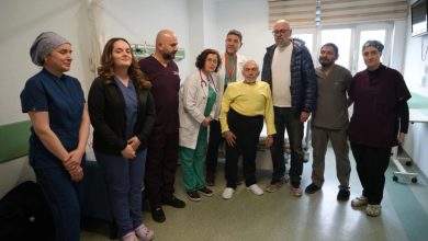 Bir hastane odasında, sağlık çalışanları ve sivil kişilerden oluşan bir grup yan yana durarak fotoğraf veriyor. Ortada ayakta duran yaşlı bir hasta dikkat çekerken, etrafında doktorlar ve sağlık personeli yer alıyor. Ortam, başarılı bir tedavi ya da ameliyat sonrası çekilmiş bir hatıra fotoğrafını andırıyor.