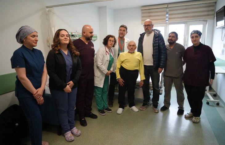 Bir hastane odasında, sağlık çalışanları ve sivil kişilerden oluşan bir grup yan yana durarak fotoğraf veriyor. Ortada ayakta duran yaşlı bir hasta dikkat çekerken, etrafında doktorlar ve sağlık personeli yer alıyor. Ortam, başarılı bir tedavi ya da ameliyat sonrası çekilmiş bir hatıra fotoğrafını andırıyor.