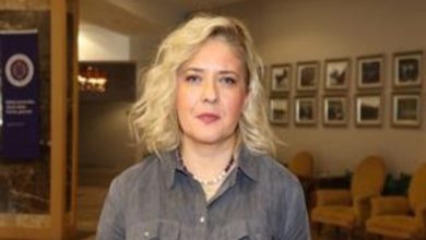 İç mekânda çekilmiş bir portre fotoğrafı. Omuz hizasında kadraja alınmış, sarı saçlı bir kadın kameraya doğrudan bakıyor. Gri tonlarında bir gömlek giymiş. Arka planda çerçeveli fotoğraflar, koltuklar ve loş bir otel ya da kurumsal mekân atmosferi görülüyor. Yüz ifadesi ciddi ve sakin.