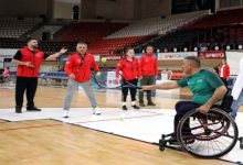 Bir spor salonunda, tekerlekli sandalyedeki bir adam raketle topa vururken, karşısında kırmızı ceketli birkaç kişi oyunu izleyip yönlendiriyor.