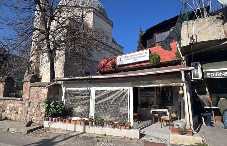 Arka planda kubbeli tarihi bir caminin yer aldığı ve önünde Edirne Bedensel Engellileri ve Ailelerini Koruma Yardımlaşma Derneği'nin binası ile sandalyelerin bulunduğu sakin bir sokak köşesi görülmektedir.