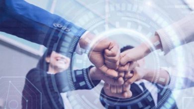 Mavi tonların hakim olduğu görselde, merkezde üst üste kenetlenmiş eller etrafında dijital halkalar ve teknoloji grafikleri yer alıyor. Arka planda ise iş birliğini simgeleyen, gülümseyen takım elbiseli profesyoneller görülüyor; genel tema birlik ve inovasyon.