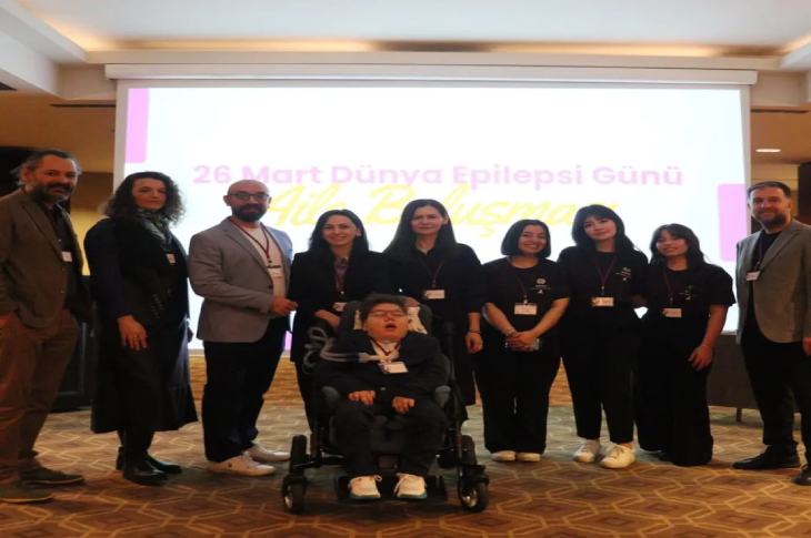 "26 Mart Dünya Epilepsi Günü Aile Buluşması" yazılı bir projeksiyon ekranının önünde, tekerlekli sandalyedeki bir çocuğun etrafında toplanmış, yaka kartlı bir grubun toplu hatıra fotoğrafı. Etkinlik salonunda çekilen karede birliktelik ve farkındalık mesajı ön planda.