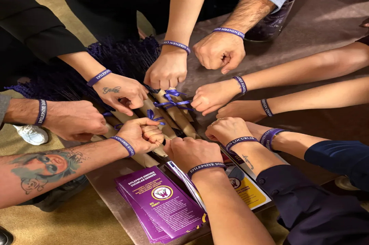 Bir masa etrafında birleşmiş, bileklerinde mor renkli "Epilepsiye Direnen Çocuklar" yazılı bileklikler bulunan ellerin kuş bakışı görüntüsü. Masada mor çiçek demetleri ve derneğe ait tanıtım broşürleri yer alıyor. Görsel, dayanışma ve farkındalık mesajı veriyor.