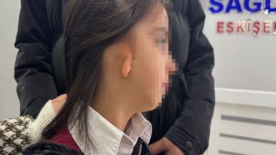 Görselde, yüzü gizlenmiş küçük bir kız çocuğu profilden görülüyor. Sağ kulak bölgesinde doğuştan gelişim farklılığı dikkat çekiyor. Yanında bir yetişkin destek amaçlı dururken, arka planda bir sağlık kurumuna ait tabela bulunuyor. Ortam, bir muayene ya da hastane sürecini çağrıştırıyor.