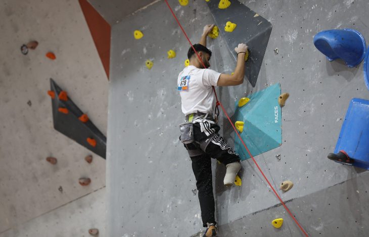 Görselde, bir sporcu yapay tırmanış duvarında yukarı doğru ilerliyor. Duvar üzerinde farklı renklerde tutamaklar bulunuyor ve sporcu emniyet ipine bağlı durumda. Odak, tırmanış sırasında sporcuların güç ve denge gerektiren hareketlerine verilmiş. Arka planda Fethiye’de düzenlenen bir para tırmanış antrenmanının atmosferi yansıyor.
