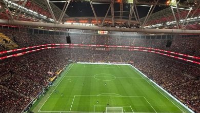 Fotoğrafta gece saatlerinde ışıklandırılmış büyük bir futbol stadyumu görülüyor. Tribünler taraftarlarla tamamen doluyken, ortada yeşil saha ve kale çizgileri net şekilde seçiliyor. Atmosfer, maç öncesi heyecanlı bir stadyum ortamını yansıtıyor.