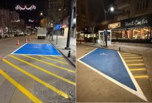 Görselde, gece saatlerinde bir şehir caddesinde engelli bireyler için ayrılmış park alanları görülüyor. Mavi zemin üzerine tekerlekli sandalye sembolü çizilmiş ve sarı çizgilerle park sınırları belirlenmiş. Yol kenarında yer alan bu alanlar, engelli sürücülerin araçlarını rahatça park edebilmesi için düzenlenmiş özel park yerlerini gösteriyor. Arkada mağazalar ve sokak aydınlatmaları da dikkat çekiyor.