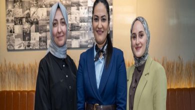 Kapalı bir mekânda yan yana duran üç kadın kameraya gülümseyerek poz veriyor. İkisi başörtülü, biri resmi bir takım elbise giymiş. Arka planda fotoğraflardan oluşan bir pano ve sade bir iç mekân dekoru görülüyor.