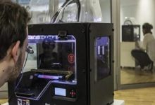 Bir laboratuvar ortamında, bir kişi masa üzerindeki 3D yazıcıyı yakından izliyor. Cihazın içinde üretim süreci devam ederken, arka planda cam bölmenin arkasında başka bir kişinin benzer bir makineyle çalıştığı görülüyor. Ortam teknik ve araştırma odaklı bir çalışma alanını andırıyor.