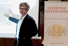 Bir sunum sırasında takım elbiseli bir uzman, projeksiyon ekranını işaret ederek anlatım yapıyor. Yanında “Lipödem Sendromu” başlıklı bir afiş yer alıyor; sahne, tıbbi bir bilgilendirme ya da konferans ortamını yansıtıyor.