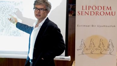 Bir sunum sırasında takım elbiseli bir uzman, projeksiyon ekranını işaret ederek anlatım yapıyor. Yanında “Lipödem Sendromu” başlıklı bir afiş yer alıyor; sahne, tıbbi bir bilgilendirme ya da konferans ortamını yansıtıyor.