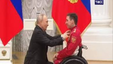 Resmi bir törende, tekerlekli sandalyedeki bir sporcuya Rusya Devlet Başkanı Vladimir Putin tarafından madalya takdim ediliyor. Arka planda bayraklar ve kürsü bulunuyor. Sahne, uluslararası bir başarıyı ve ödüllendirme anını yansıtıyor.