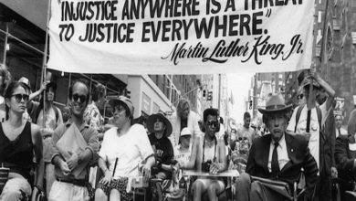 Şehrin merkezinde, yüksek binaların arasında ilerleyen bir grup engelli aktivist, tekerlekli sandalyeleriyle güçlü ve kararlı bir protesto yürüyüşü gerçekleştiriyor. Kalabalığın önünde taşınan pankartta Martin Luther King Jr.’ın şu sözü yer alıyor: “Injustice anywhere is a threat to justice everywhere.” (Herhangi bir yerdeki adaletsizlik, her yerdeki adalet için tehdittir.) Bu ifade yalnızca bir alıntı değil; eşitlik, erişilebilirlik ve hak temelli mücadelenin evrensel çağrısını simgeleyen güçlü bir hatırlatma niteliği taşıyor. Yürüyüş, dayanışmanın görünür hâle geldiği, adalet talebinin kamusal alanda kararlılıkla dile getirildiği bir hak savunuculuğu örneği olarak öne çıkıyor.