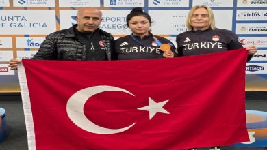 Görselde bir sporcu kazandığı bronz madalya ile Türkiye bayrağını iki kişiyle birlikte tutarak poz veriyor. Üç kişi “Türkiye” yazılı eşofmanlarla şampiyona alanında gururla fotoğraf çektiriyor. Arkada organizasyonun sponsor panosu yer alıyor.