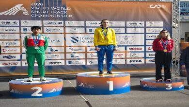 Fotoğrafta bir spor müsabakasının ödül töreni görülüyor. Üç sporcu madalya kürsüsünde yer alıyor. Ortada birinci olan sporcu en yüksek basamakta altın madalyayla dururken, solda ikinci olan sporcu gümüş madalya ile ikinci basamakta bulunuyor. Sağda ise üçüncü basamakta bronz madalya kazanan bir sporcu yer alıyor. Arka planda “2026 Virtus World Short Track Championship” yazılı sponsor panosu bulunuyor ve sporcular boyunlarında madalyalarıyla kürsüde poz veriyor.