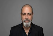 Gri arka plan önünde çekilmiş bir portre fotoğrafı görülüyor. Orta yaşlı bir erkek doğrudan kameraya bakıyor. Kısa kesilmiş saçları, belirgin kaşları ve gri-beyaz karışımı sakalı var. Siyah bir ceket ve koyu renk gömlek giymiş; kulağında küçük halka küpeler ve boynunda ince bir zincir bulunuyor. Yüz ifadesi ciddi ve sakin.