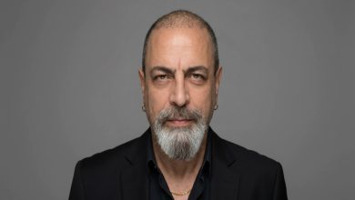 Gri arka plan önünde çekilmiş bir portre fotoğrafı görülüyor. Orta yaşlı bir erkek doğrudan kameraya bakıyor. Kısa kesilmiş saçları, belirgin kaşları ve gri-beyaz karışımı sakalı var. Siyah bir ceket ve koyu renk gömlek giymiş; kulağında küçük halka küpeler ve boynunda ince bir zincir bulunuyor. Yüz ifadesi ciddi ve sakin.