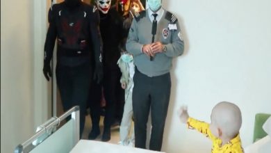 Bir hastane odasında, yatağın yanında duran küçük bir çocuk kapıya doğru elini uzatıyor. Kapıdan içeri, süper kahraman ve palyaço (Joker benzeri) kostümleri giymiş kişiler girerken, önde bir görevli maske takarak ayakta duruyor. Ortamda sürpriz bir ziyaret ve neşeli bir an hissediliyor.