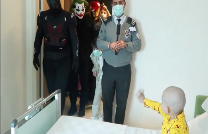 Bir hastane odasında, yatağın yanında duran küçük bir çocuk kapıya doğru elini uzatıyor. Kapıdan içeri, süper kahraman ve palyaço (Joker benzeri) kostümleri giymiş kişiler girerken, önde bir görevli maske takarak ayakta duruyor. Ortamda sürpriz bir ziyaret ve neşeli bir an hissediliyor.