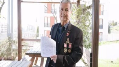 Görselde, açık alandaki bir oturma bölümünde ayakta duran bir erkek görülüyor. Üzerinde madalyaların yer aldığı koyu renk bir ceket bulunuyor. Elinde bir belge tutarak kameraya doğru gösteriyor. Arka planda binalar, ağaçlar ve ahşap pergola yapısı yer alıyor.