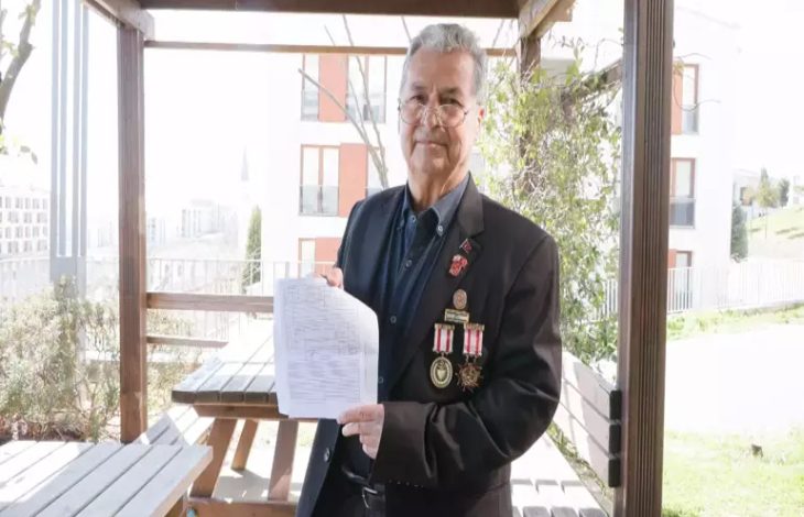 Görselde, açık alandaki bir oturma bölümünde ayakta duran bir erkek görülüyor. Üzerinde madalyaların yer aldığı koyu renk bir ceket bulunuyor. Elinde bir belge tutarak kameraya doğru gösteriyor. Arka planda binalar, ağaçlar ve ahşap pergola yapısı yer alıyor.