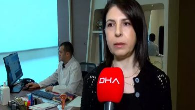 Bir sağlık merkezinde röportaj veren bir kadın, elinde kırmızı mikrofonla kameraya konuşuyor. Arka planda bir doktor bilgisayar başında çalışıyor; masa üzerinde tıbbi materyaller ve bir rahim modeli bulunuyor. Ortam klinik ve düzenli görünüyor.