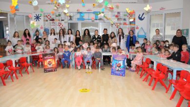 Bir anaokulu sınıfında çok sayıda çocuk ve öğretmen toplu fotoğraf çektiriyor. Sınıf renkli süslemeler, küçük sandalyeler ve oyuncaklarla dolu. Ön tarafta ağız ve diş sağlığına dikkat çeken afişler bulunuyor; ortam bir eğitim etkinliğini yansıtıyor.