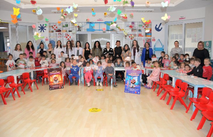 Bir anaokulu sınıfında çok sayıda çocuk ve öğretmen toplu fotoğraf çektiriyor. Sınıf renkli süslemeler, küçük sandalyeler ve oyuncaklarla dolu. Ön tarafta ağız ve diş sağlığına dikkat çeken afişler bulunuyor; ortam bir eğitim etkinliğini yansıtıyor.