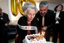 Yaşlı bir kadın, üzerinde “İyi ki doğdun” yazılı kuşakla, üzerinde fotoğraf bulunan doğum günü pastasındaki “100” mumlarını üflerken görülüyor. Yanında bir erkek ona destek olurken, arka planda altın renkli “100” balonlar ve kutlamaya katılan kişiler yer alıyor. Atmosfer sıcak ve duygusal bir 100. yaş kutlamasını yansıtıyor.