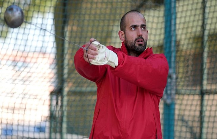 Kırmızı eşofmanlı bir sporcu, çekiç atma kafesinde atışa hazırlanırken görülüyor. Eldivenli elleriyle çekiç topunu savururken odaklanmış bir ifadeye sahip. Görseldeki kişi, işitme engelli milli atlet Muhammed Çakır.