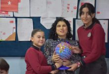 Fotoğrafta bir sınıf ortamında ortada oturan bir öğretmen ile yanında iki öğrenci görülüyor. Öğrenciler öğretmenle birlikte renkli bir dünya küresi tutuyor. Arka planda duvara asılmış öğrenci çizimleri ve Türk bayrağı bulunan sınıf panosu yer alıyor.