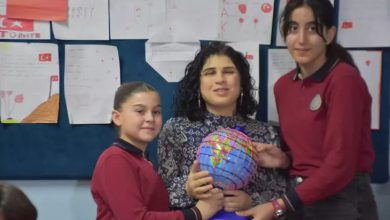 Fotoğrafta bir sınıf ortamında ortada oturan bir öğretmen ile yanında iki öğrenci görülüyor. Öğrenciler öğretmenle birlikte renkli bir dünya küresi tutuyor. Arka planda duvara asılmış öğrenci çizimleri ve Türk bayrağı bulunan sınıf panosu yer alıyor.