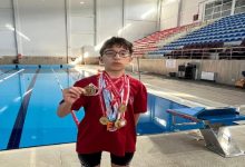 Kapalı bir yüzme havuzunda çekilmiş fotoğrafta, gözlüklü bir çocuk sporcu havuz kenarında ayakta duruyor. Boynunda çok sayıda madalya asılı, elinde ise bir madalyayı kameraya doğru tutuyor. Kırmızı tişört ve yüzme şortu giymiş. Arka planda havuz, kulvarlar ve tribün koltukları görülüyor; sahne net biçimde sportif bir başarı anını yansıtıyor.