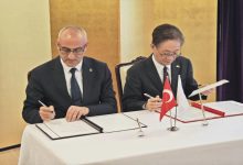 Bir masada yan yana oturan iki resmi giyimli kişi, önlerindeki belgeleri imzalıyor. Masanın üzerinde Türk bayrağı ve Japon bayrağı küçük masa bayrakları olarak yer alıyor. Ortam, resmi bir anlaşma veya protokol imza törenini andırıyor.