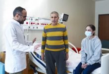 Fotoğrafta bir hastane odasında doktor, hasta ve yanında bulunan genç bir kadın görülüyor. Hasta ayakta dururken doktor onunla konuşuyor, genç kadın ise yatak kenarında oturarak durumu dikkatle izliyor. Ortam, organ nakli süreciyle ilgili bir hastane kontrolünü yansıtıyor.