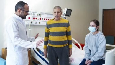 Fotoğrafta bir hastane odasında doktor, hasta ve yanında bulunan genç bir kadın görülüyor. Hasta ayakta dururken doktor onunla konuşuyor, genç kadın ise yatak kenarında oturarak durumu dikkatle izliyor. Ortam, organ nakli süreciyle ilgili bir hastane kontrolünü yansıtıyor.