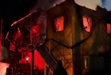 Gece saatlerinde iki katlı bir evin alevler içinde kaldığı görülüyor. Pencerelerden yoğun turuncu ışık yayılırken çatının bir kısmı çökmüş, merdiven ve dış cephe is içinde. Ön planda bir kişi yangını izlerken ortamda yoğun duman ve acil durum hissi hakim.