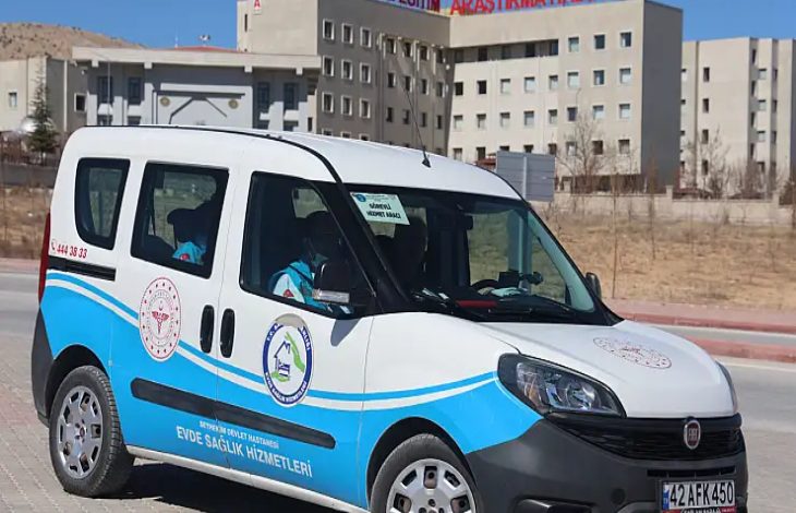 Görselde, bir hastanenin önünde park etmiş “Evde Sağlık Hizmetleri”ne ait resmi bir hizmet aracı görülüyor. Üzerinde sağlık birimlerinin amblemleri ve iletişim bilgileri yer alan araç, açık havada gündüz saatlerinde çekilmiş. Arka planda büyük bir eğitim ve araştırma hastanesi binası bulunuyor.